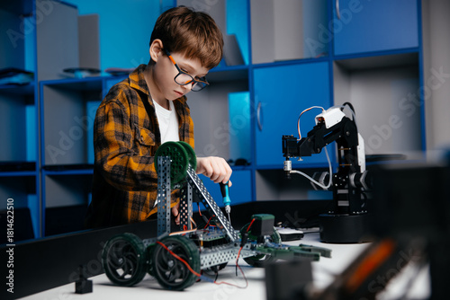 Young boy with glasses is assembling a robotic model in a modern workshop, surrounded by tools and equipment, showcasing creativity and engineering skills in action