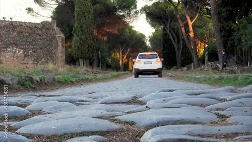 Tableau sur toile Ancient Roman road made of large cobblestones with a modern car driving away, bl