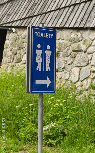 Public toilet direction sign with green grass and wall background providing clear guidance. Blue restroom sign with male and female icons standing near stone building in outdoor area during daylight.