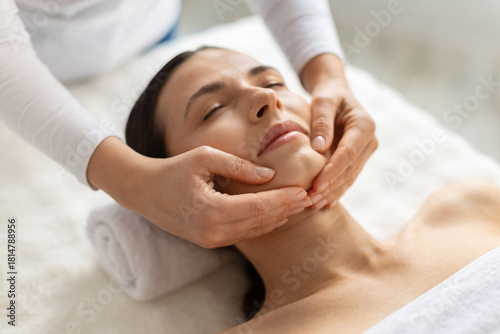 Young beautiful woman, female client receiving gentle chin massage at spa. Calm expression, warm light and skilled hands highlight beauty, skincare care, wellness and a peaceful lifestyle atmosphere