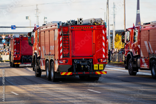 Red fire trucks in organized convoy showing rear doors, reflective graphics, tool storage and ladder mounts as they travel along marked street with crowd in background