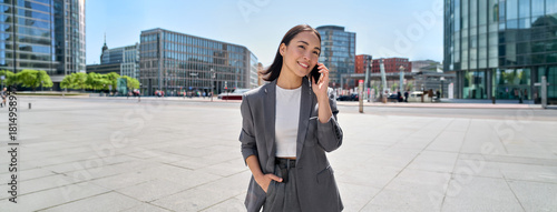Young Asian successful businesswoman leader wearing suit standing in big city talking on mobile phone. Smiling woman making business call on cell walking on busy downtown street outdoors
