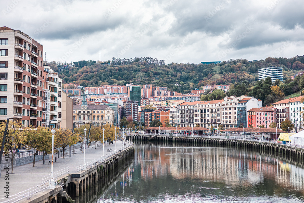 Obraz premium wide view of the Bilbao riverfront, with colorful buildings reflected in the calm water. The city rises up a wooded hillside under a dramatic, cloudy sky, showcasing urban and natural elements.
