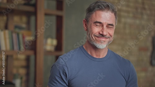 Portrait of happy confident casual middle aged man at home in loft room. Fit and healthy active senior male with grey hair looking at camera, smiling. Books in background, smart older guy.