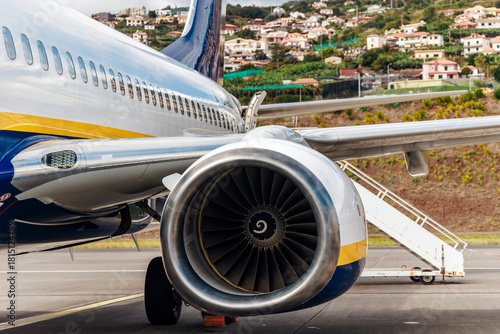 Commercial airplane parked on runway, showcasing detailed engine design and sleek fuselage, with residential buildings on hillside in the background, highlighting aviation technology