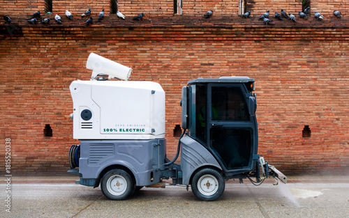 electric street cleaning vehicle spraying water as it moves past the old brick wall of Tha Phae gate in Chiang mai, with pigeons perched above during daytime
