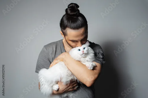 Fototapeta Studio portrait of young man with white cat in arms while kissing the pet on grey background.