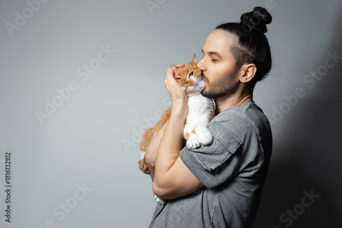 Studio portrait of young man with red cat in arms while kissing the pet on grey background.