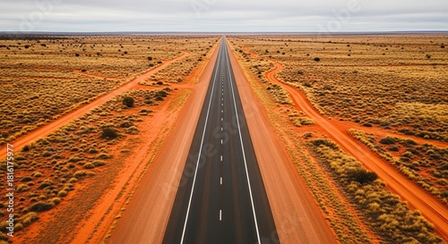 High angle aerial view of a long, straight asphalt highway stretching to the horizon through a vast, remote outback landscape with red earth and sparse vegetation under an overcast sky