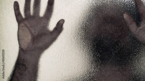 Blurred human figure presses hands against frosted glass door creating eerie horror tension. Distorted silhouette suggests fear and confinement. For suspense, danger, and psychological concepts.
