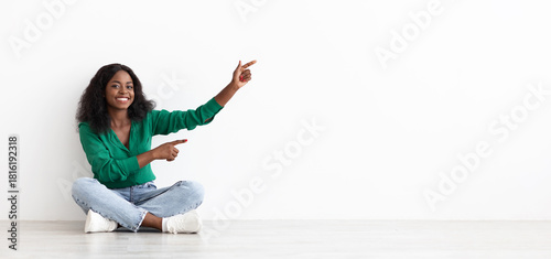 A young woman with curly black hair sits on the floor, wearing a green top and jeans. She smiles and points excitedly to the left, creating a friendly atmosphere in a bright room.