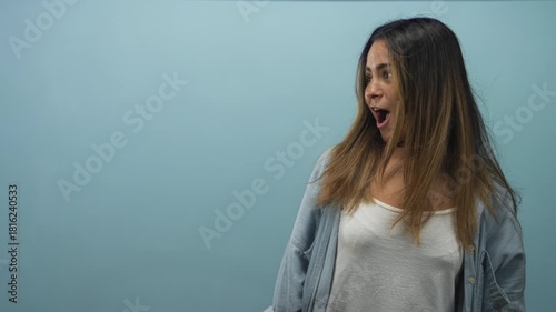 Hispanic teenage woman with open mouth and surprised expression looking sideways in pale blue studio setting; surprise wonder curiosity.