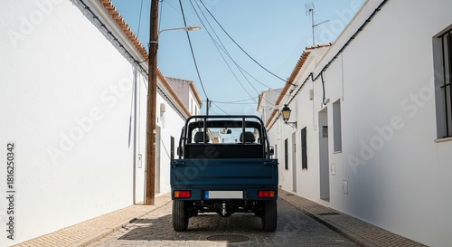 Fototapeta Naklejka Na Ścianę i Meble -  A small blue utility truck is parked on a narrow cobblestone street between traditional whitewashed buildings in a quiet European village on a sunny day