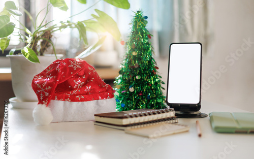 working on christmas holidays concept: close up view of mobile phone, red hat, notebook and christmas tree decoration on table