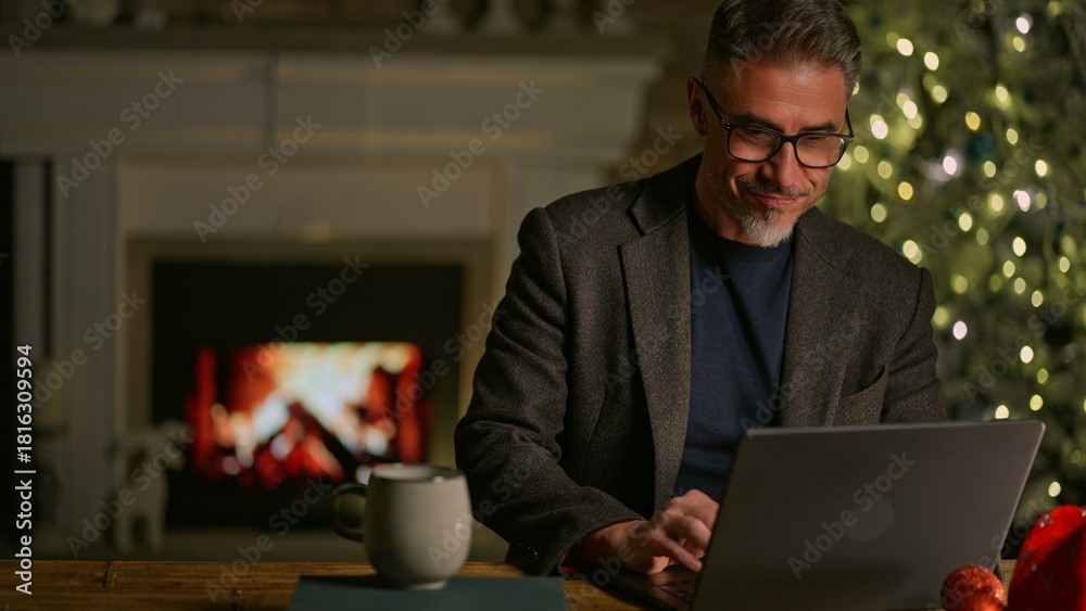 Naklejka premium Happy smiling mid adult man using laptop computer at home, sitting at desk in Christmas decorated cosy room. Fireplace and Christmas tree in background. Businessman shopping online searching for gift.
