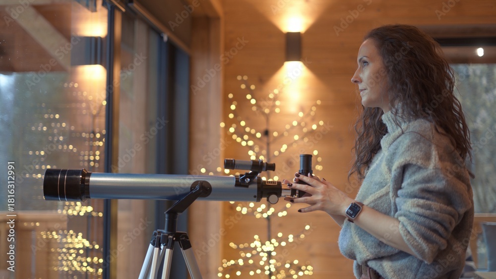 Fototapeta premium Middle aged woman at home looking out window with telescope. Female in turtleneck pullover standing in living room of wooden cabin with Christmas lights. Autumn or winter indoor clothing.