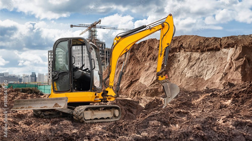 Obraz premium Mini excavator is actively digging at a busy construction site, with towering buildings and a crane visible under a partly cloudy sky