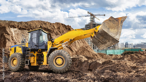 A large yellow wheeled bulldozer or loader is digging and relocating earth at a busy construction site surrounded by buildings and cranes under a dramatic sky
