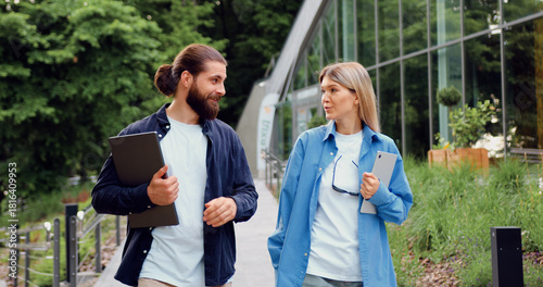 Two business professionals, man with laptop and woman with tablet, wearing casual office attire, walking outside, while engaged in a work-related conversation, showcasing teamwork
