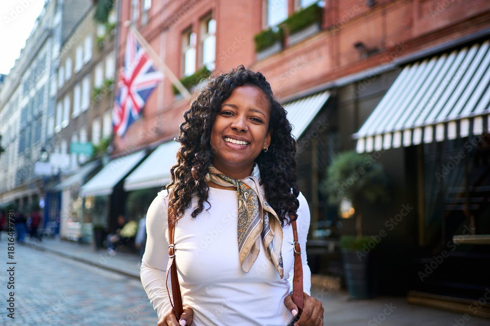 Fototapeta premium Cheerful millennial woman holds her backpack straps looking at camera on a lively London street, with Union Jack and striped awnings behind her, capturing a bright travel moment with confident energy
