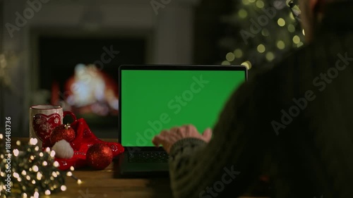 Man shopping online on Internet with laptop computer at Christmas. Christmas tree and lights in cosy winter home with fireplace. Blank green screen for web shop offer and holiday advertisement.