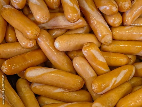 A dense, close-up pile of cooked, glossy, light brown cocktail sausages or small hot dogs.