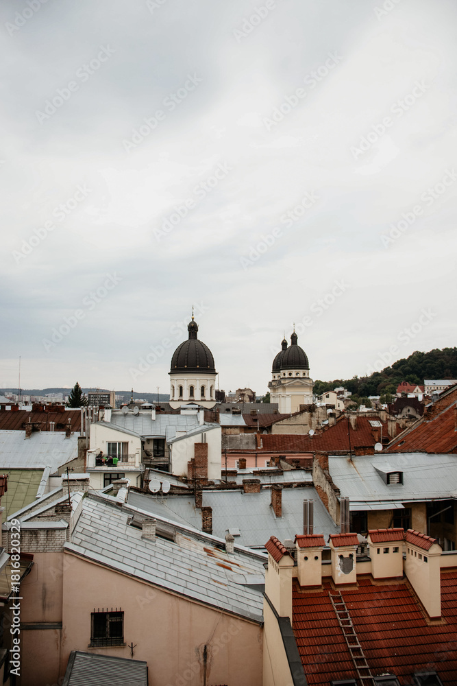 Fototapeta premium An overcast rooftop view of Lviv's historic center, featuring a stately, weathered old church facade with a red tile roof surrounded by light-colored European buildings.