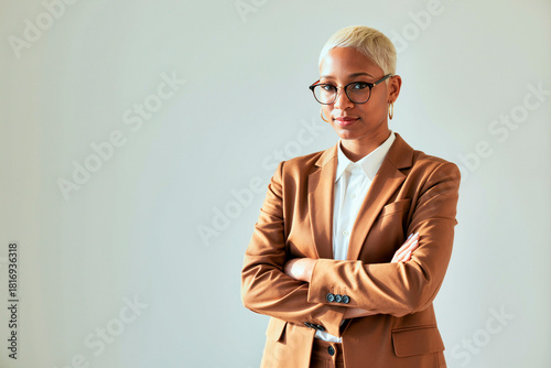 Portrait of young black woman with short blond hair and glasses standing with arms crossed in office setting, looking confidently at camera, business context