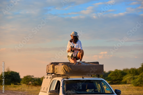 People on Safari theme: Solo Woman on the rooftop tent observing savanna for animals. at Golden Hour, Self-Drive Safari, Nxai Pan, Botswana.