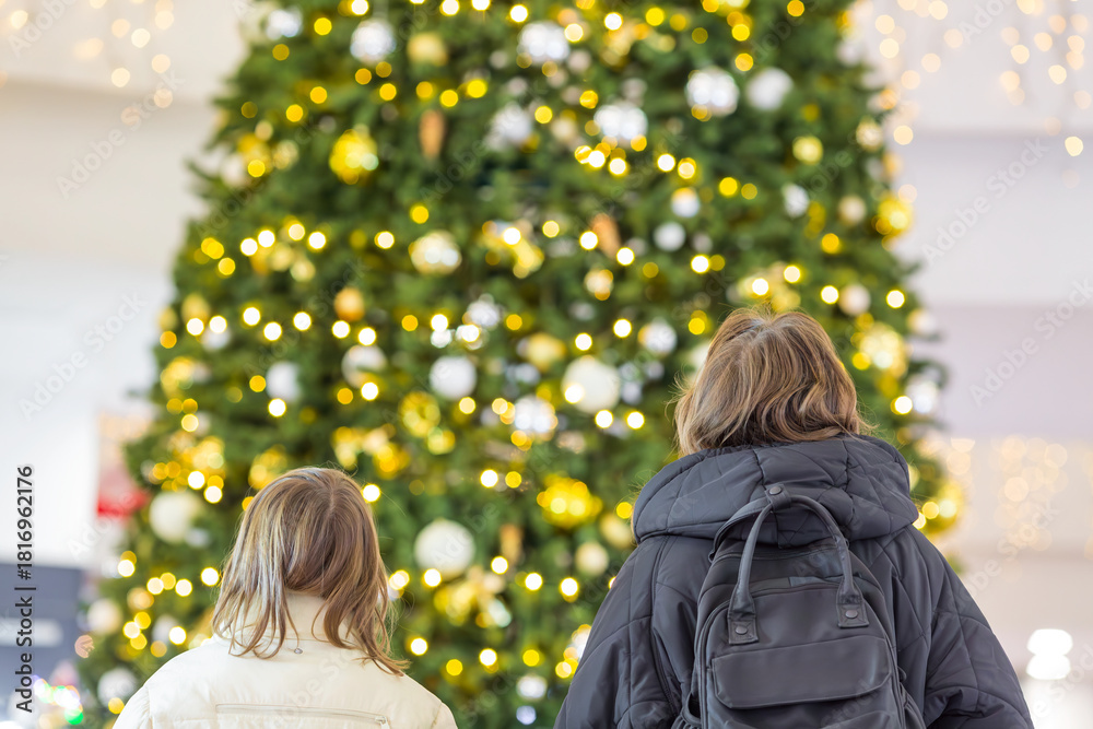 Obraz premium Young two beautiful girls sisters standing in front of decorated Christmas tree with lights in shopping mall. Photo from behind. Christmas fairytale