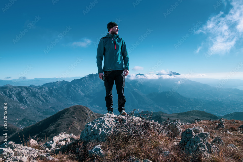 Obraz premium A lone hiker sits on a rocky ridge, quietly taking in the vast mountain landscape beneath a clear, teal-tinted sky.