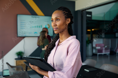 Thoughtful black young businesswoman with tablet in office with colleagues in background