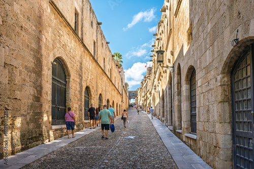 Fototapeta Naklejka Na Ścianę i Meble -  Tourists walk the cobblestone pedestrian Street of the Knights, a 14th century street medieval street in the old town of Rhodes, Greece.