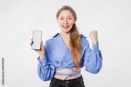 Happy young woman looking at camera holding phone in studio on white background casual clothes celebrating good news social media communication concept