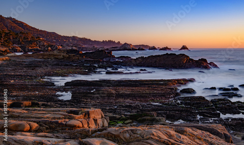 Coastal long-exposure landscape with a dramatic sunset over the water, where the sun meets the ocean waves near the rocks, offering a serene nature view. 