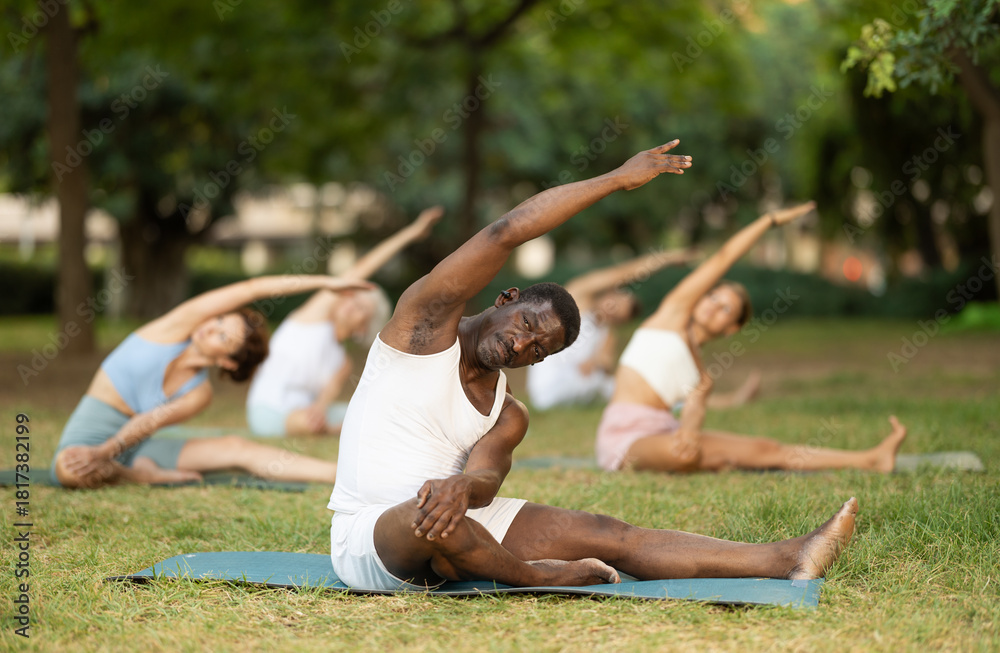 Naklejka premium Sporty African American in comfortable white sportswear practicing hatha yoga during group class on green lawn in summer park, sitting on mat and doing stretching in Parivrtta Janu Sirsasana asana