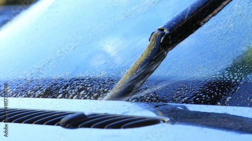 Close-up view of a car windshield being cleaned with a wiper, showcasing water droplets and dirt removal, camera pans across the scene highlighting the action flow