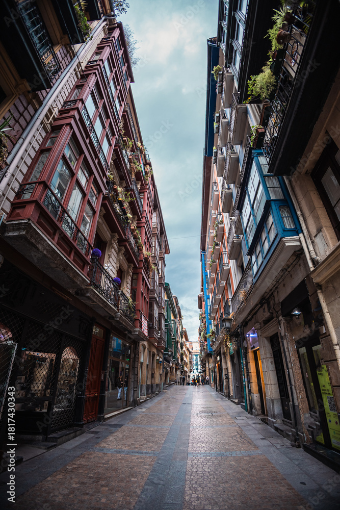 Obraz premium A dramatic low-angle shot up a narrow street in Bilbao's Casco Viejo, showing tall, richly detailed buildings with iron balconies and glassed-in bay windows meeting under a cloudy sky.
