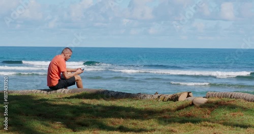 A man wears an orange shirt and shorts as he relaxes on a log by the ocean. He is focused on his smartphone. Copy space. 