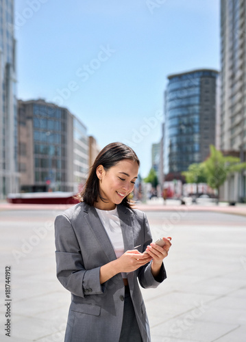 Young happy Asian business woman wearing suit holding mobile phone standing in big city on busy street, smiling lady worker using smartphone for texting, cellphone apps for work outside, vertical
