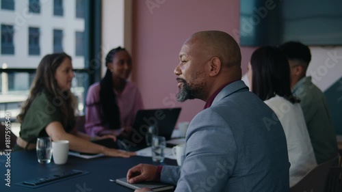 Smiling mature black businessman looking at camera while sitting with colleagues in boardroom at office
