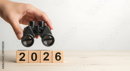 Hand holding a pair of black binoculars over wooden blocks spelling out the year 2026, symbolizing future vision, planning, and looking ahead to the next years goals and prospect .