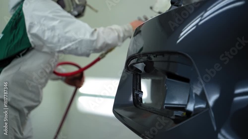 Auto technician spray gloss lacquer on car bumper inside clean, ventilated paint booth. Wearing protective coveralls and a respirator using a high precision spray gun.