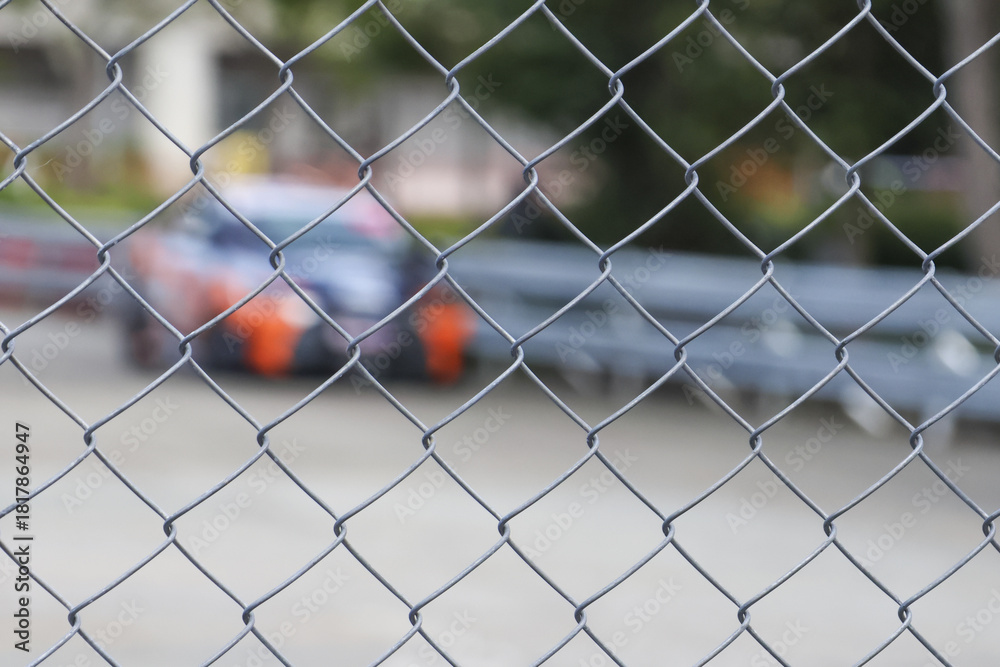 Fototapeta premium Selective focus on metal chain link fence creating an abstract wire mesh pattern. blurred race car in background evokes feeling of security and separation