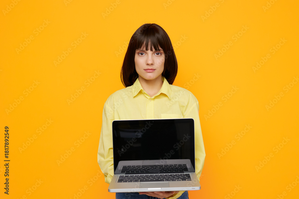 Naklejka premium Young woman stands upfront with a laptop extended toward the camera against a bright orange background, presenting a clean, modern tech inspired portrait.