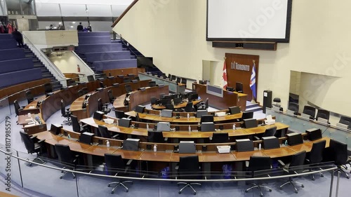 An elevated, wide-angle view from the public gallery of the unoccupied Toronto City Council Chamber, highlighting the circular arrangement of desks and screen