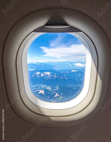 Aerial view of mountainous terrain and cloud-filled sky as seen through an airplane window. Sunlight casts shadows on frame