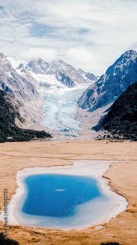 A vast alpine landscape featuring a bright blue lake in the foreground, surrounded by dry, golden grass. In the background, a massive glacier flows down from sn