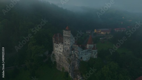 Misty sunrise aerial orbit around Bran Castle in Transylvania, Romania, revealing the medieval fortress and surrounding village in panoramic view