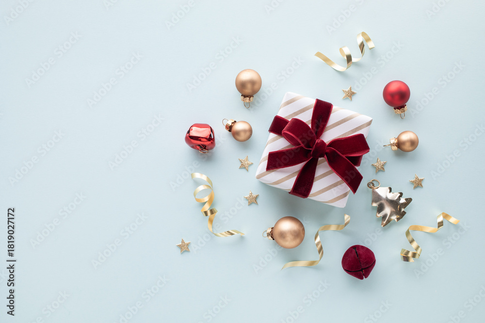 Obraz premium Gift box golden wrapped with red velvet ribbon on light blue backdrop decorations festive bauble and stars for Christmas and New Year greeting card
